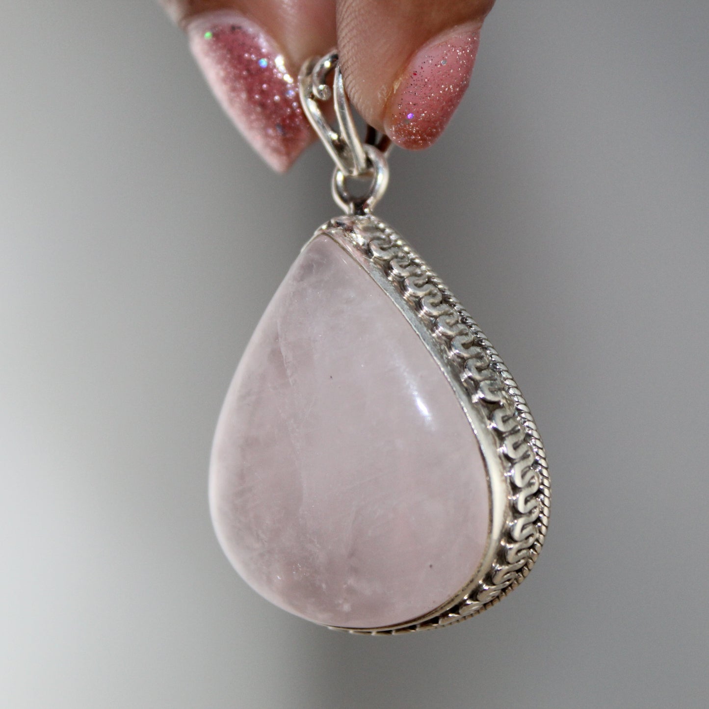 Teardrop-shaped pink stone pendant with silver frame held by a hand against a gray background