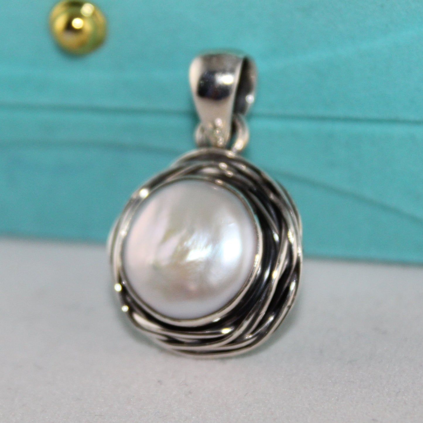 Pearl pendant in silver setting on a light gray surface with a turquoise box in the background.