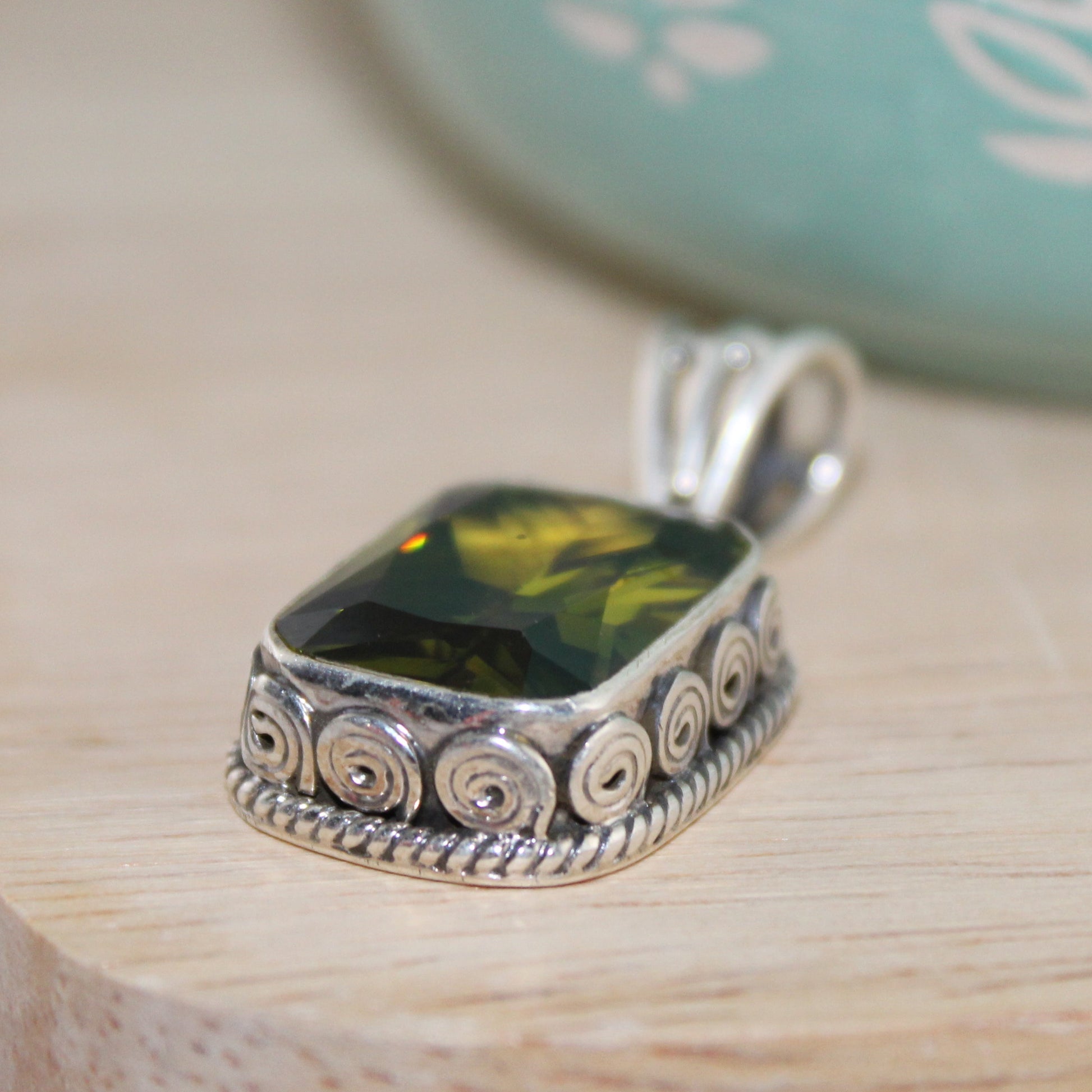 Silver pendant with a green gemstone on a wooden surface with a light blue decorative object in the background.
