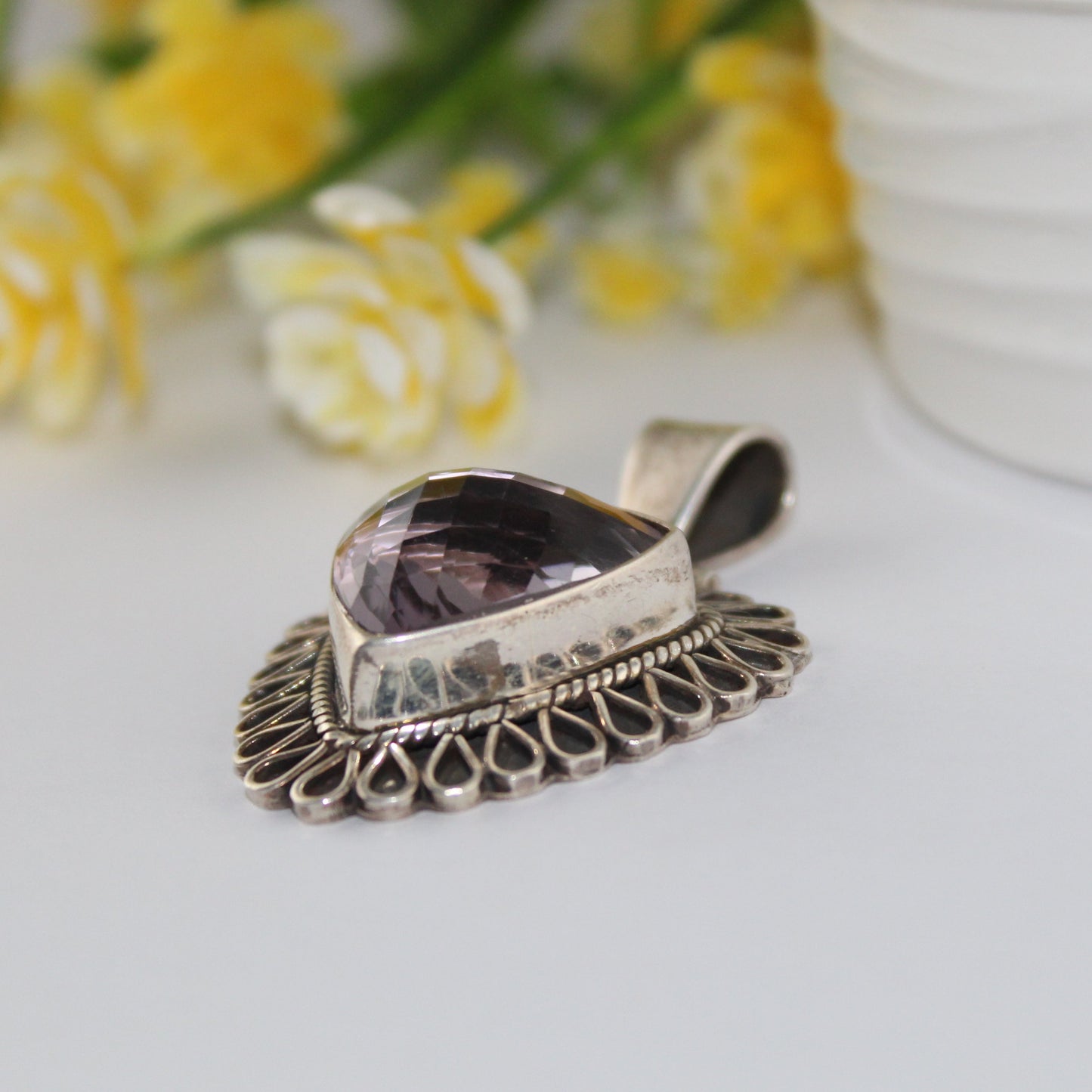 Silver pendant with a purple amethyst stone on a white surface with yellow flowers in the background